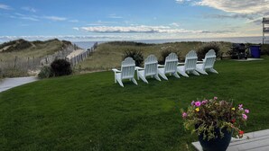 A grassy area with white Adirondack chairs arranged in a row, overlooking a beach and dunes. - Oceanview Condo w/ Fireplace & Patio (Dennis Port)
