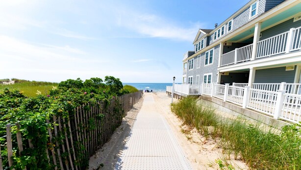 A beachfront walkway with a wooden fence on one side and a building with balconies on the other. - Oceanview Condo w/ Fireplace & Patio (Dennis Port)