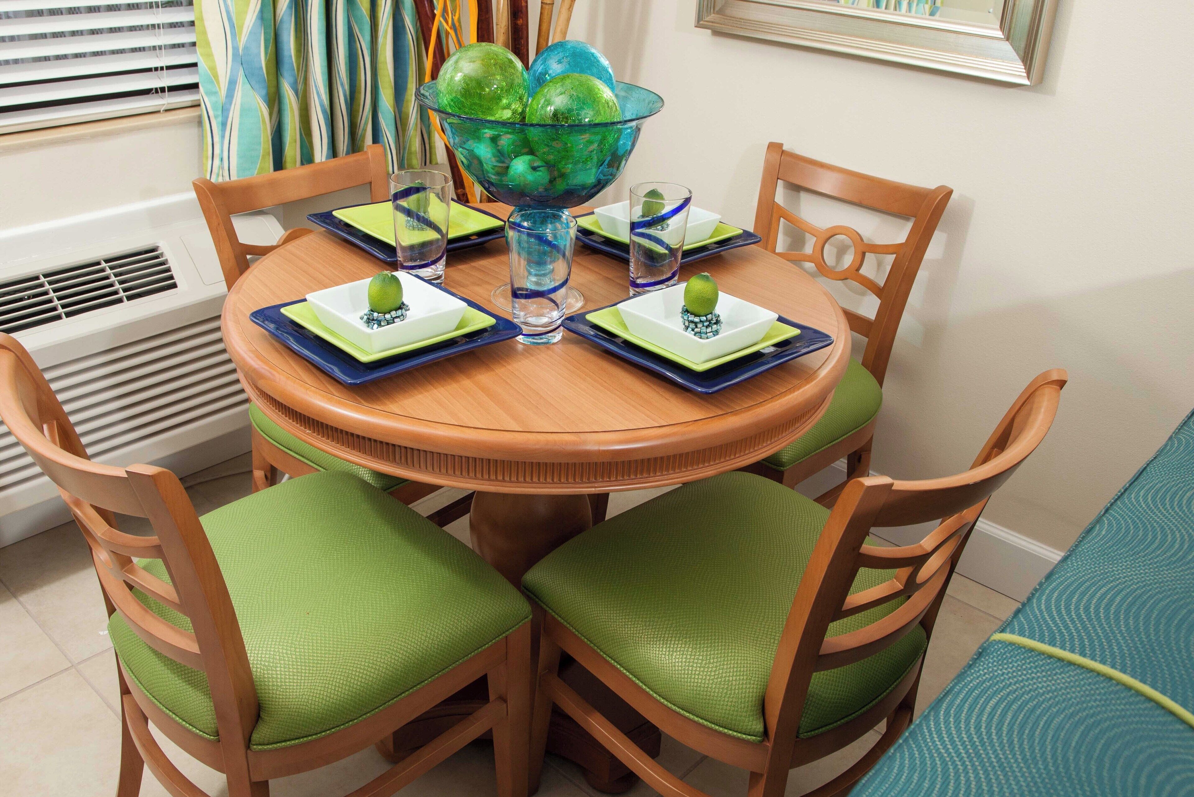 Une table de salle à manger ronde en bois, avec des chaises à coussins verts, un saladier en verre contenant des objets décoratifs verts, et des sets de table bleus et verts.