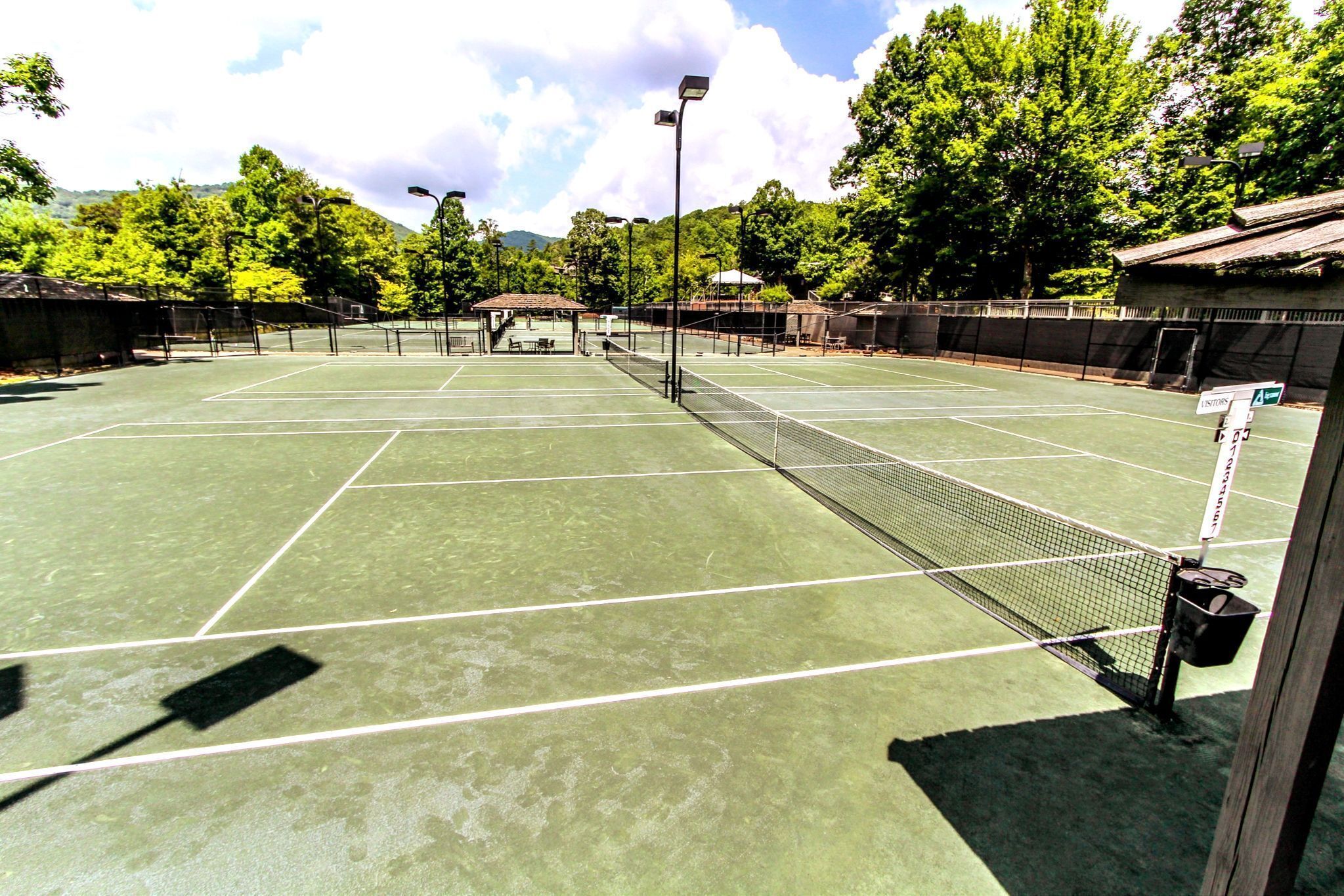 A tennis court with multiple courts, a covered seating area, and surrounding trees.