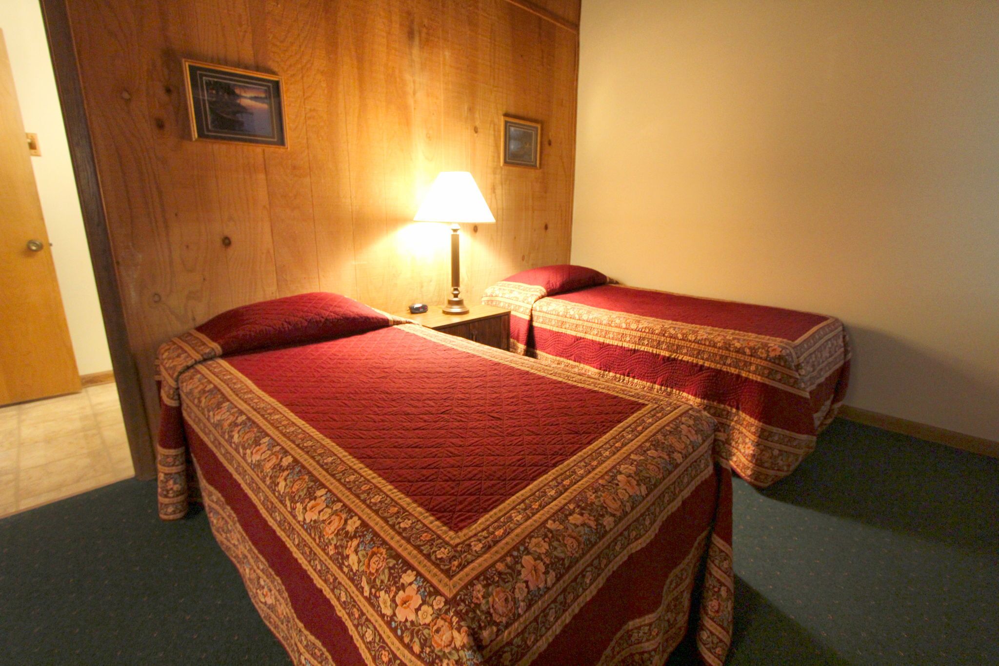 A room with two beds, a wooden wall, a lamp, and a framed picture.