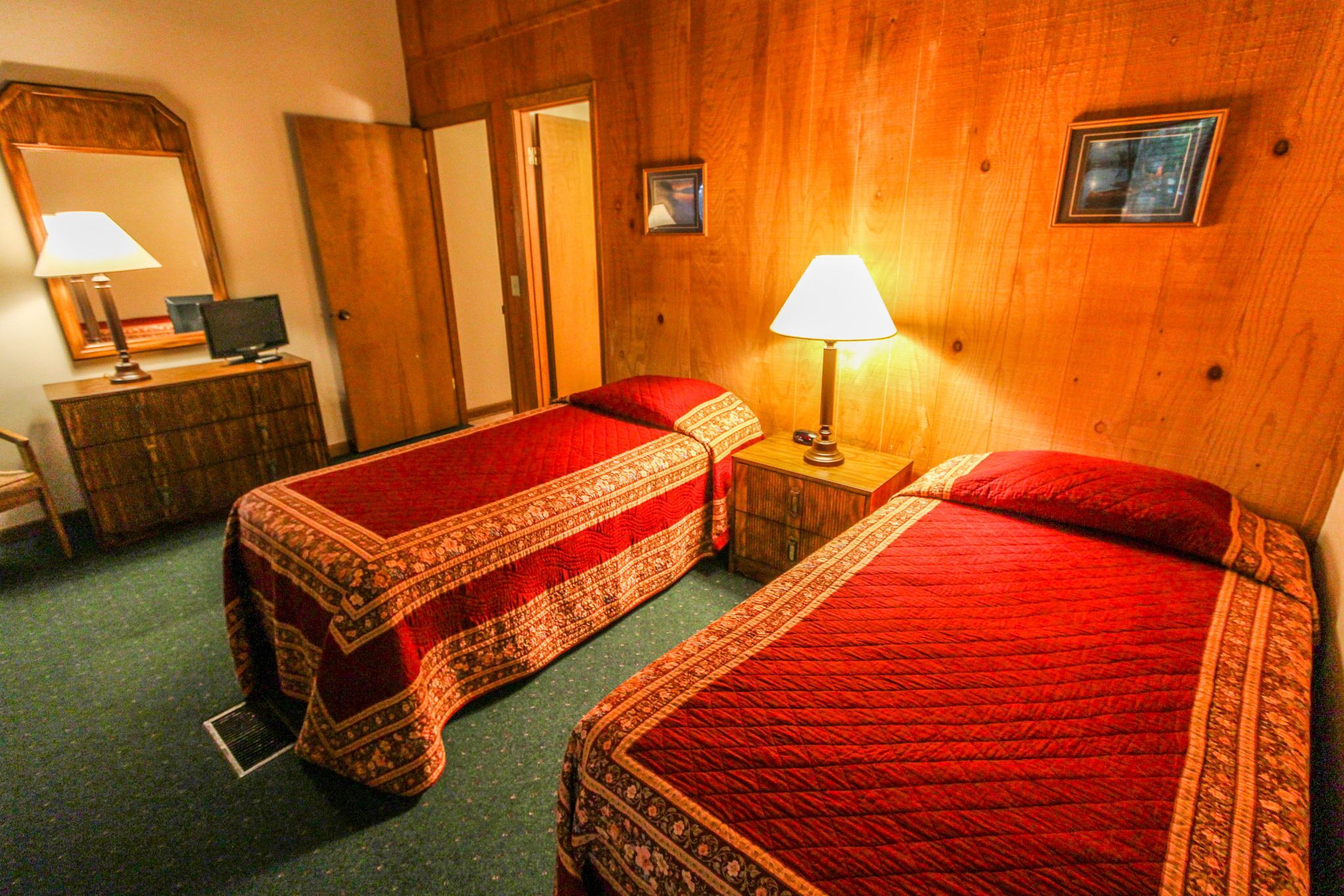 A hotel room with two single beds, a wooden headboard, a bedside table with a lamp, a dresser, a mirror, and a door.