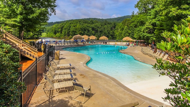 A swimming pool with a curved design, surrounded by lounge chairs and umbrellas, set against a backdrop of lush greenery and hills. - Mountain Condo w/ Deck & Fireplace (Marble Hill)