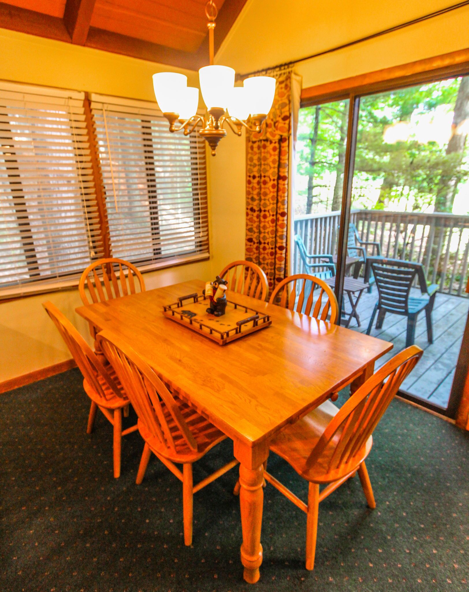 Une table à manger en bois, avec six chaises, un lustre et une vue sur une terrasse accessible par une porte vitrée.
