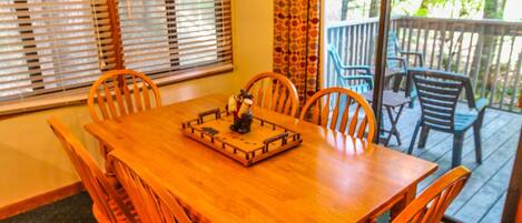 Une table à manger en bois, avec six chaises, un lustre et une vue sur une terrasse accessible par une porte vitrée.