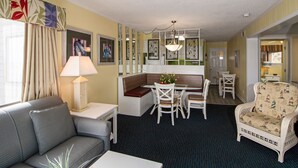 A hotel room with a sofa, a dining table with chairs, a small kitchen area, and a decorative chair.
