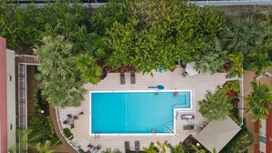 Vista aérea de una piscina rodeada de palmeras y sillas de descanso, con un deck empedrado y un área de estar cubierta.