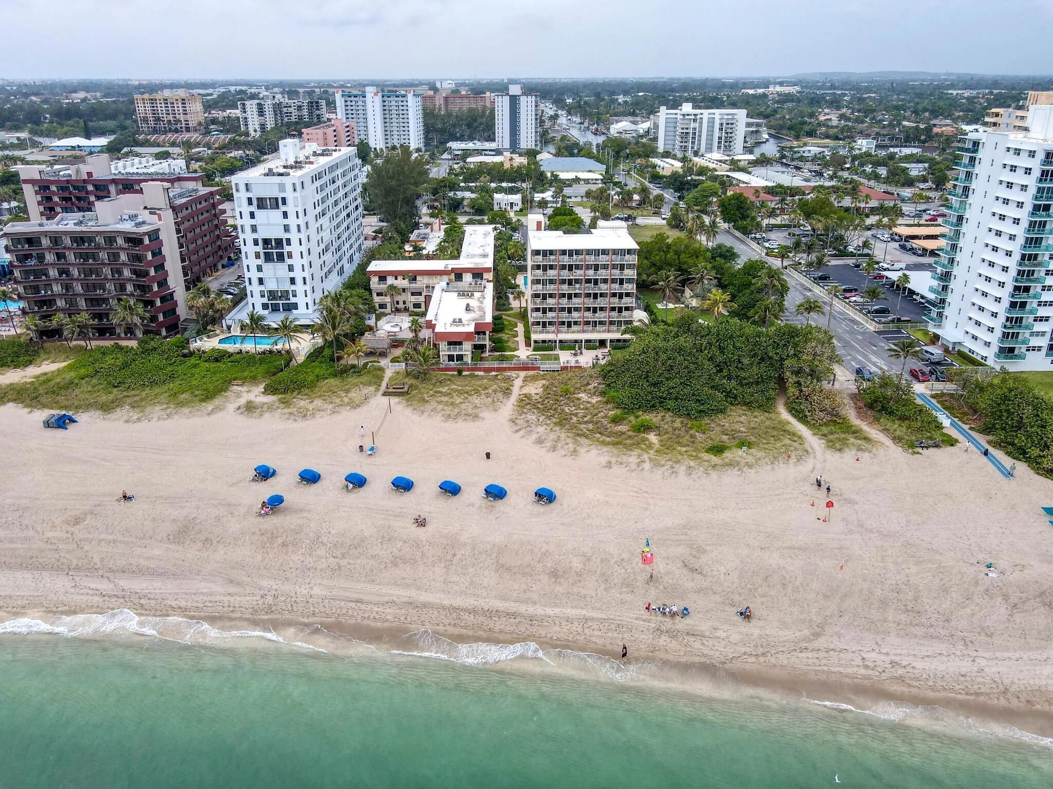 An aerial view of a beach with several buildings in the background, including high-rise hotels and residential structures.