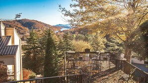 A wooden deck with chairs and a table, surrounded by trees and a mountain view. - Mountain Getaway | Pools, Trails & More! (Lake Lure)