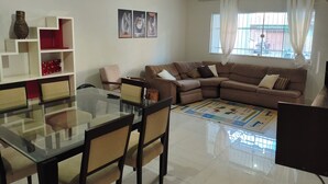 A living room with a glass dining table, beige chairs, a brown sectional sofa, and a red 'LOVE' sign on a white shelf.