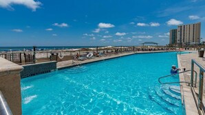 A clear blue pool with lounge chairs and umbrellas, a view of the ocean, and a tall building in the background. - Phoenix X 803 | Beachfront | Next to Flora-Bama | Pools & Hot Tubs | My Beach Getaways (Orange Beach)