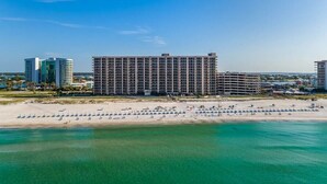 A large hotel with multiple floors, a beach with rows of umbrellas, and a clear view of the ocean. - Phoenix X 803 | Beachfront | Next to Flora-Bama | Pools & Hot Tubs | My Beach Getaways (Orange Beach)