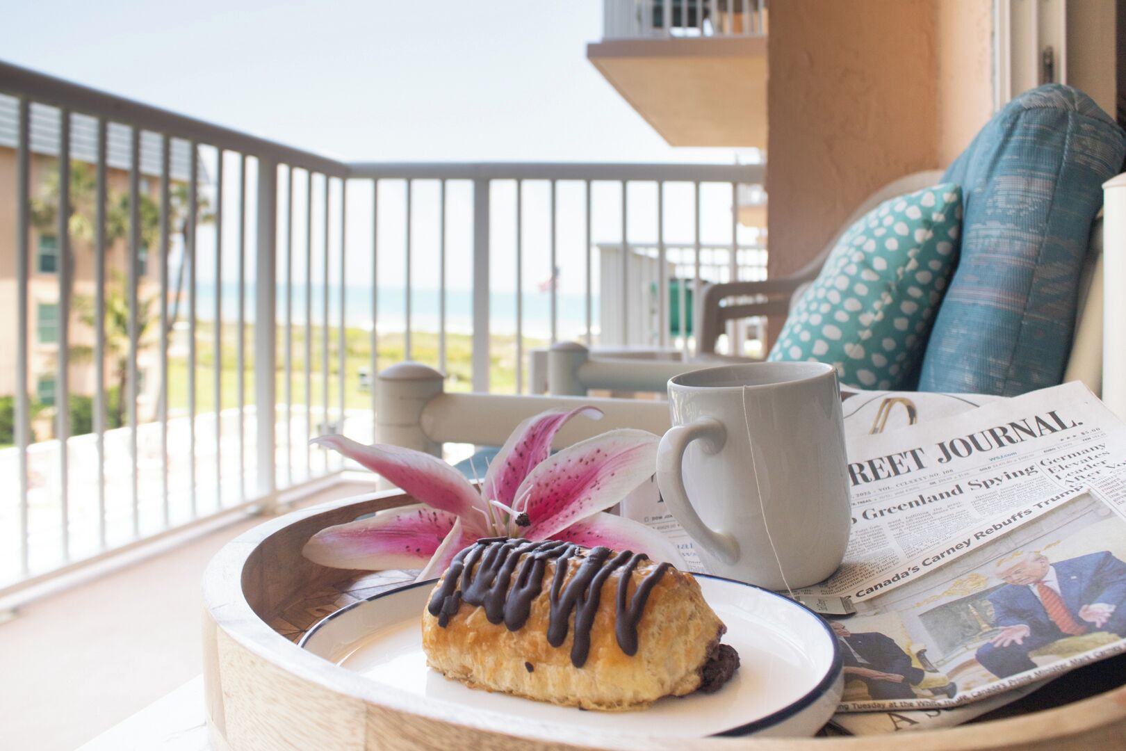 Un plateau de petit-déjeuner avec une pâtisserie, une tasse et un journal, posé sur un balcon donnant sur un immeuble et de la verdure.