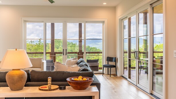 A spacious living room with a large sliding glass door leading to a deck, a comfortable sofa, a wooden table with a bowl of fruit, and a ceiling fan. - Calico Haven at BRMC | Hot Tub, Views, Pool (Boone)