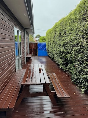 A wooden deck with a picnic table and benches, adjacent to a house with a blue tarp and a neatly trimmed hedge.