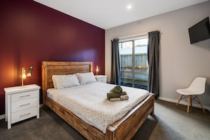 A bedroom with a wooden bed, white nightstands, a white chair, a TV on the wall, and a window with curtains.