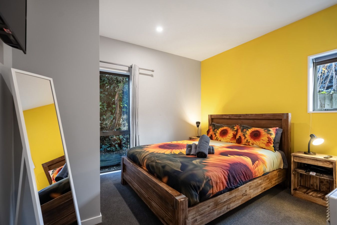 A bedroom with a wooden bed, a nightstand, a mirror, and a window with a view of greenery.