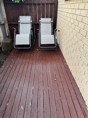 Dos sillones de exterior con estructura metálica y asientos de malla, ubicados en una terraza de madera con un muro de ladrillo al fondo.