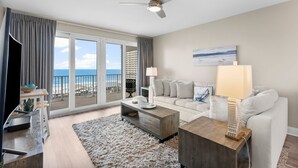 A modern living room with a sofa, coffee table, and a ceiling fan. A large window offers a view of the ocean and a high-rise building.