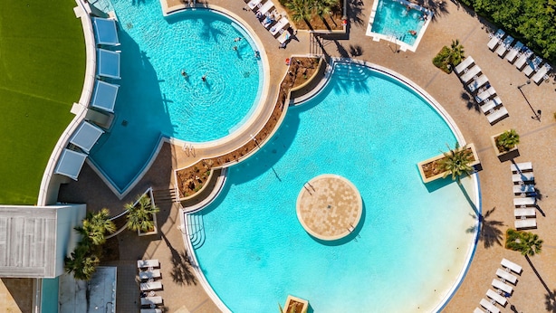 A large, multi-pool resort with a central circular pool, surrounded by sun loungers and palm trees.