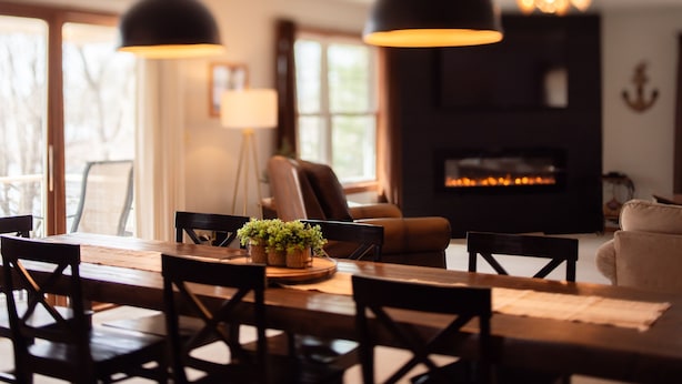 Een eetruimte met een houten tafel, stoelen en hangende lampen. Achterin is een open haard te zien.