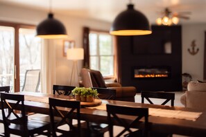 Un coin repas avec une table en bois, des chaises et des lampes suspendues. On aperçoit une cheminée au fond.