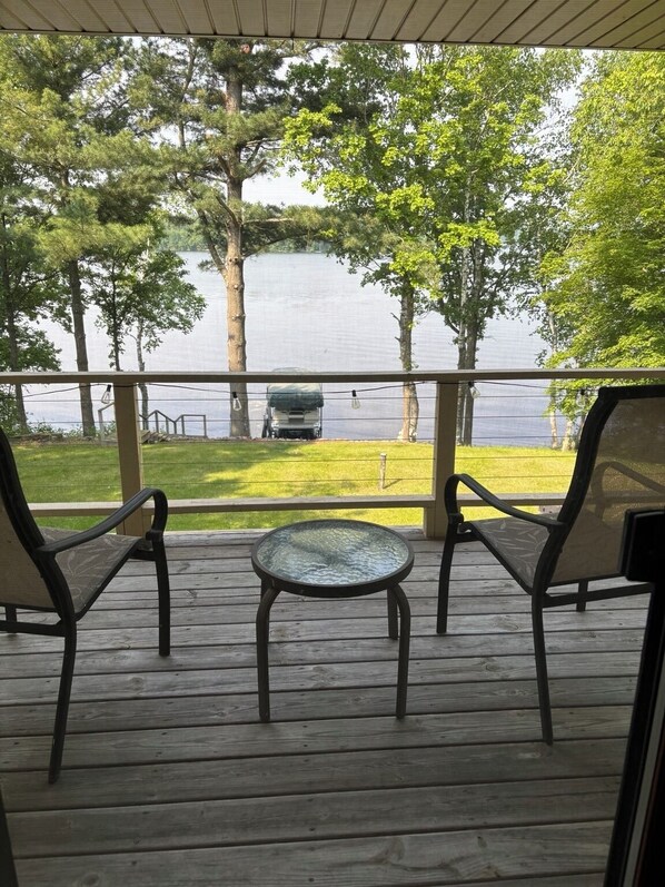 Une terrasse en bois avec deux chaises et une petite table, donnant sur un lac bordé d’arbres et sur lequel se trouve un bateau.