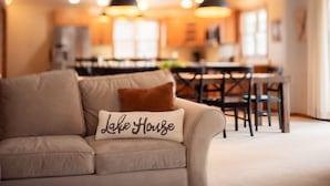 Een woonkamer met een beige bank, een kussen met de tekst 'Lake House', een eethoek met een houten tafel en stoelen, en hanglampen.