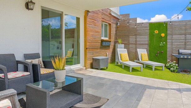 A modern outdoor patio with wicker furniture, a glass-top table, and a green lawn area with lounge chairs. - Pool & Canal-View Townhouse | Miami Oasis + BBQ (Miami)