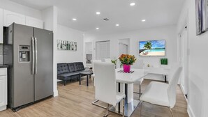 A modern kitchen with a stainless steel refrigerator, a dining area with white chairs and a table, and a living area with a sofa and a TV displaying a beach scene. - Pool & Canal-View Townhouse | Miami Oasis + BBQ (Miami)