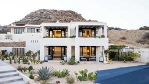 A modern two-story house with a desert garden, featuring a staircase, outdoor seating, and a view of a rocky hill.