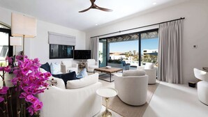 A modern living room with white furniture, a ceiling fan, a flat-screen TV, and a large window with curtains.