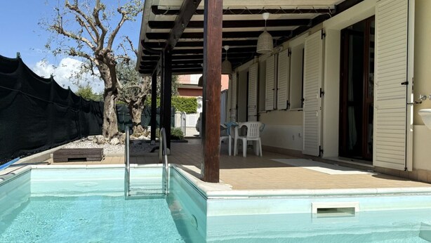 Een zwembad met een pergola erboven, een overdekt terras en een gebouw met witte luiken.