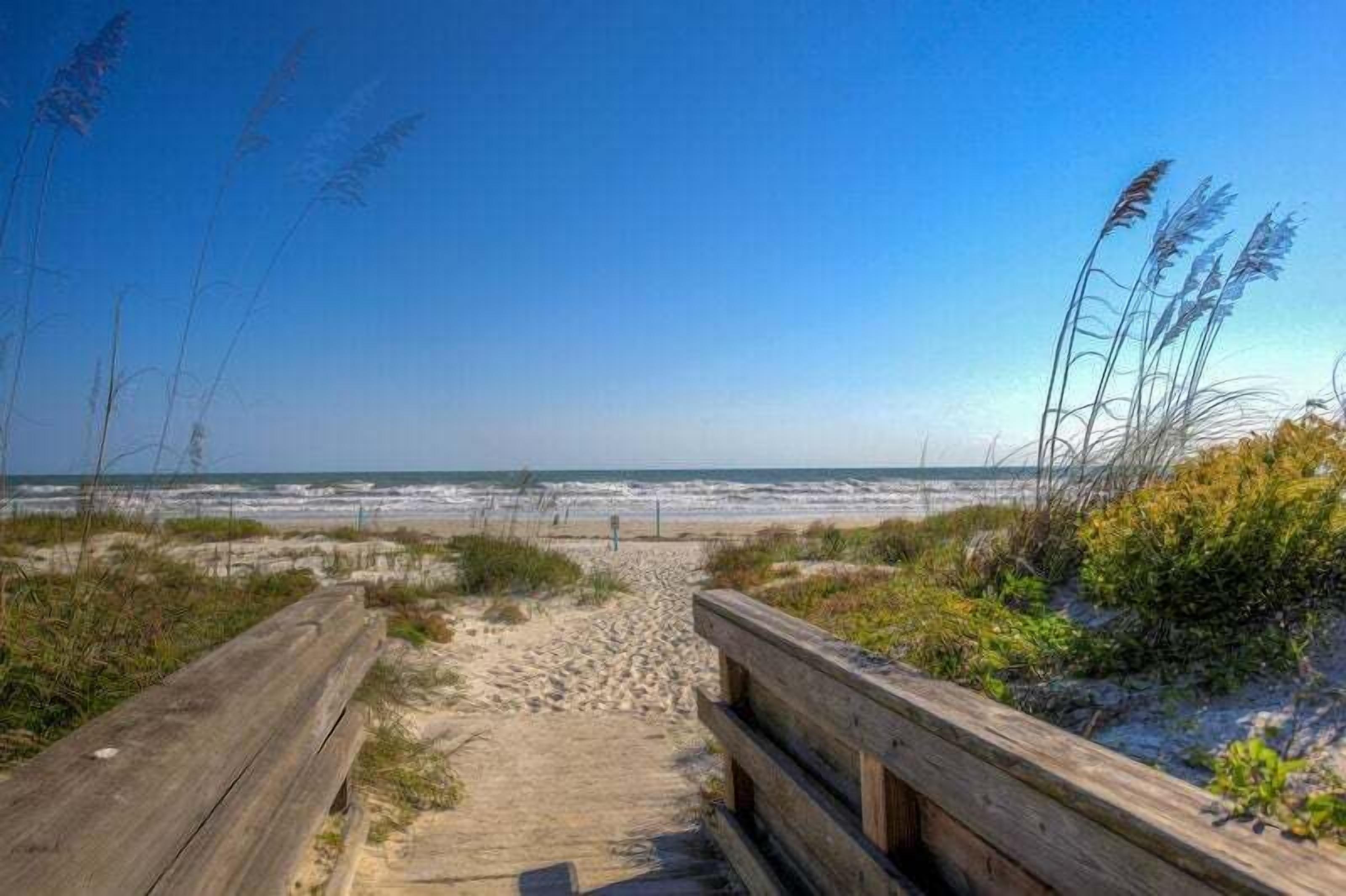 Un paseo marítimo de madera que lleva a una playa con dunas de arena y pastos marinos.