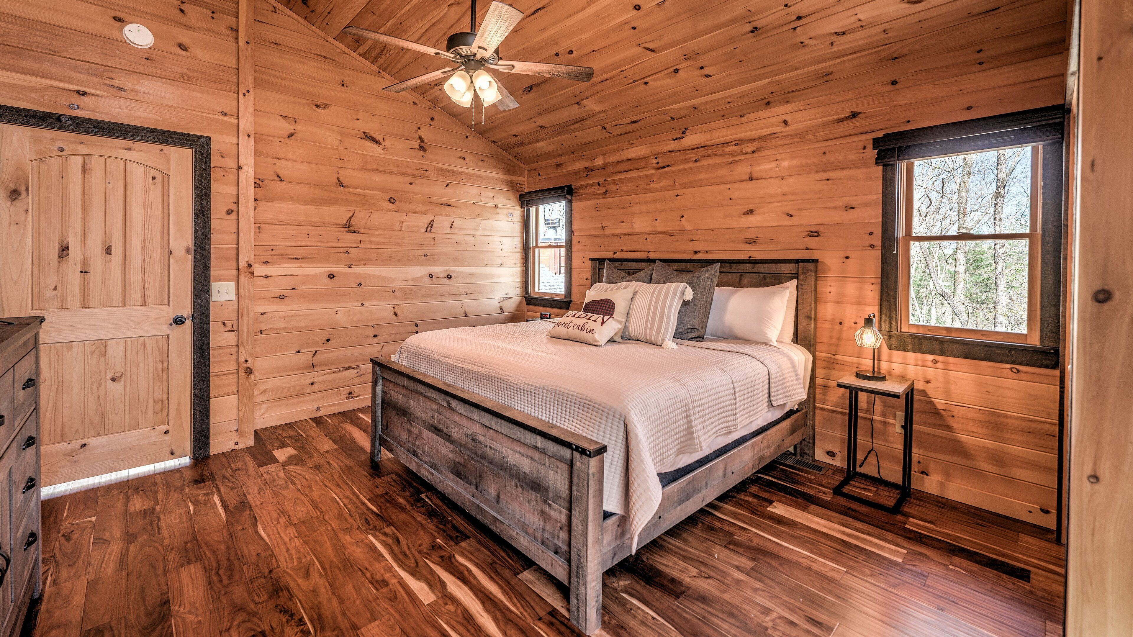 Quarto de cabana de madeira com uma cama, mesa de cabeceira, cômoda e ventilador de teto.