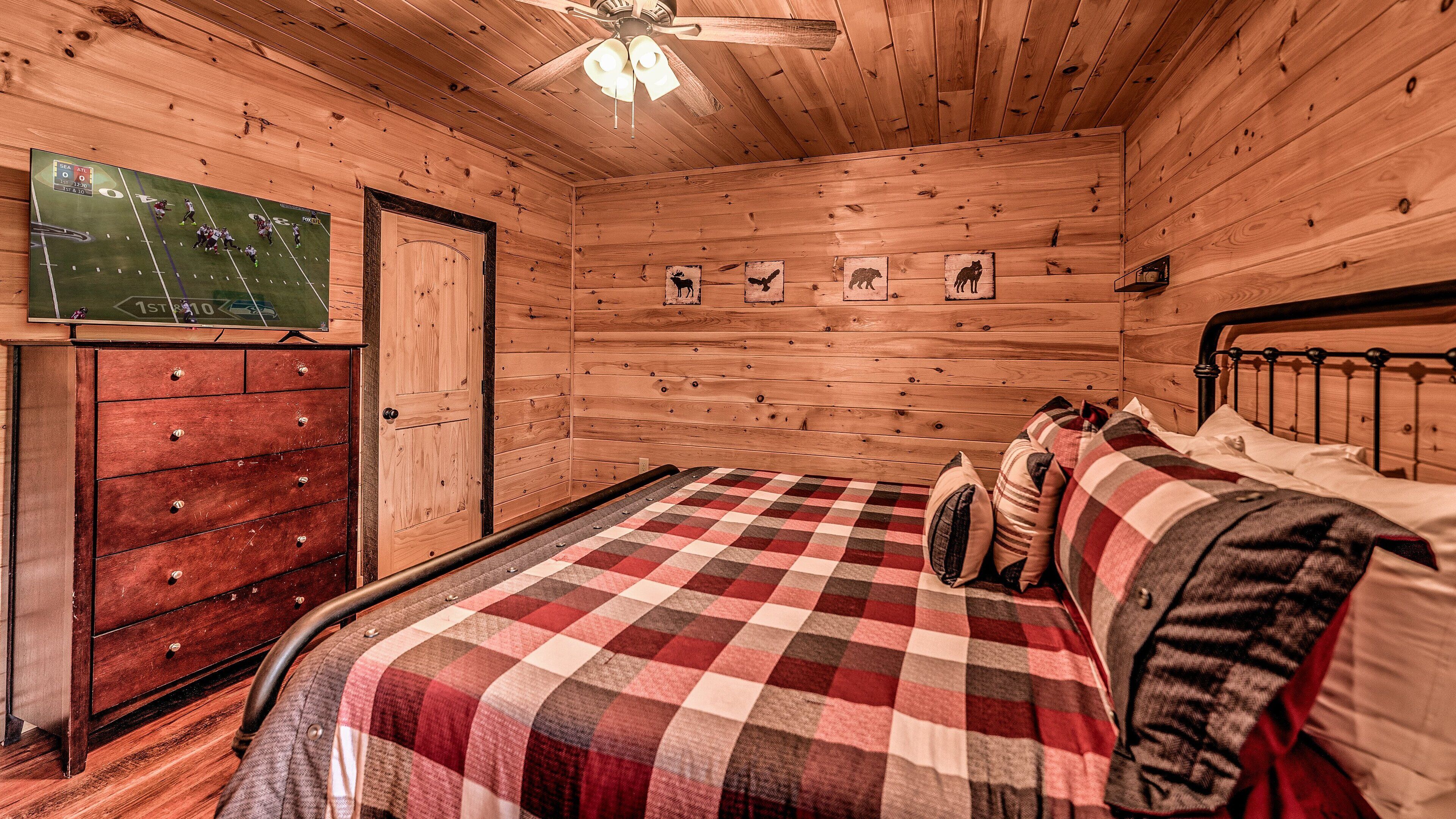A wooden cabin bedroom with a bed, a dresser, a ceiling fan, and a wall-mounted TV displaying a football game.