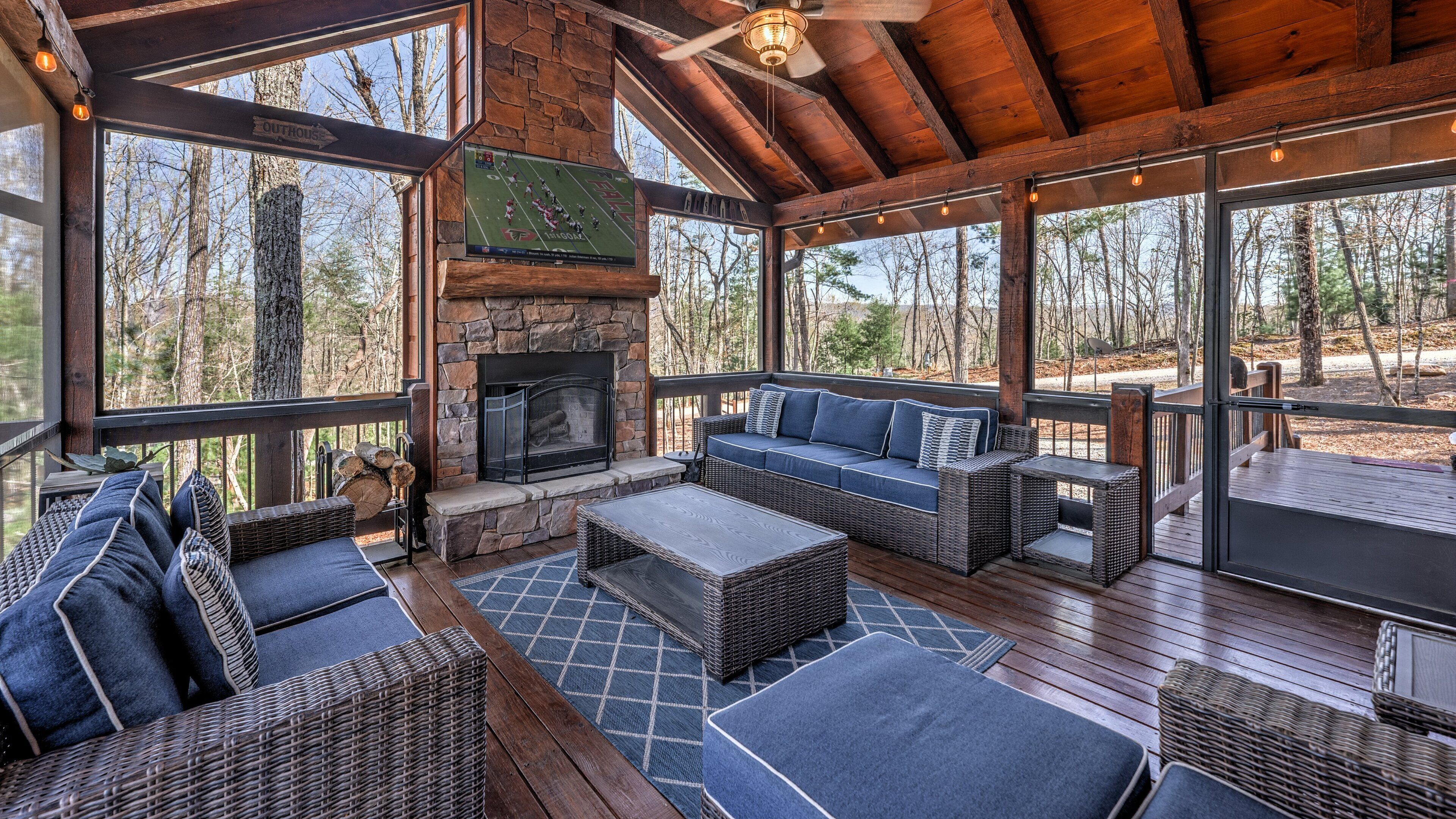 A covered outdoor seating area with a stone fireplace, wicker furniture, and large windows offering forest views.