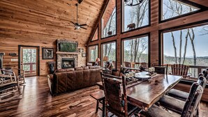 Interior espacioso de una cabaña con chimenea, pisos de madera, una gran mesa de comedor y múltiples ventanas que ofrecen vistas al bosque circundante.