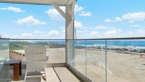 Un balcón con baranda de vidrio que da a una playa, una mesa de madera y una silla blanca.