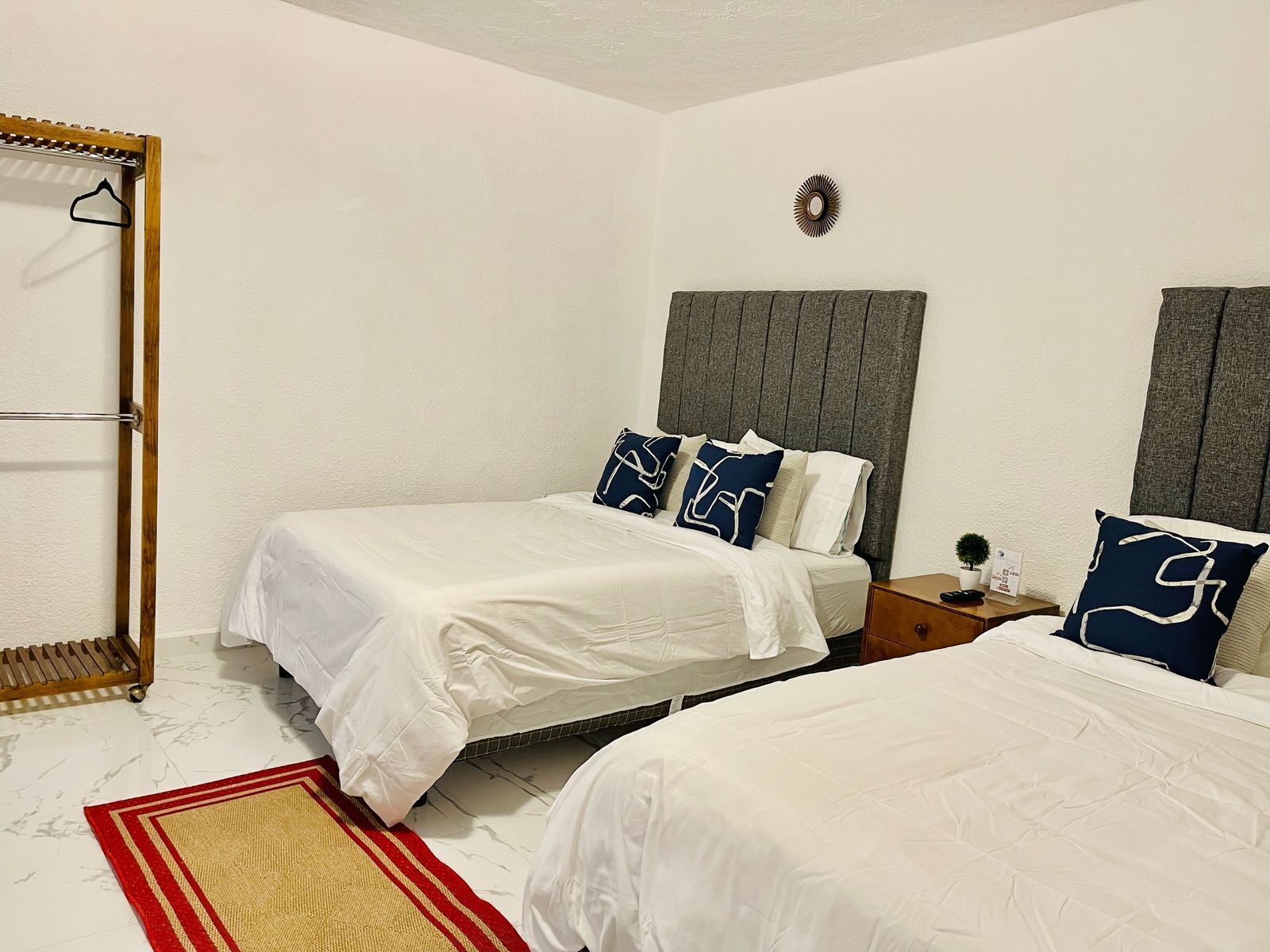 A hotel room with two single beds, a wooden wardrobe, a small table with a plant, and a red and beige rug.