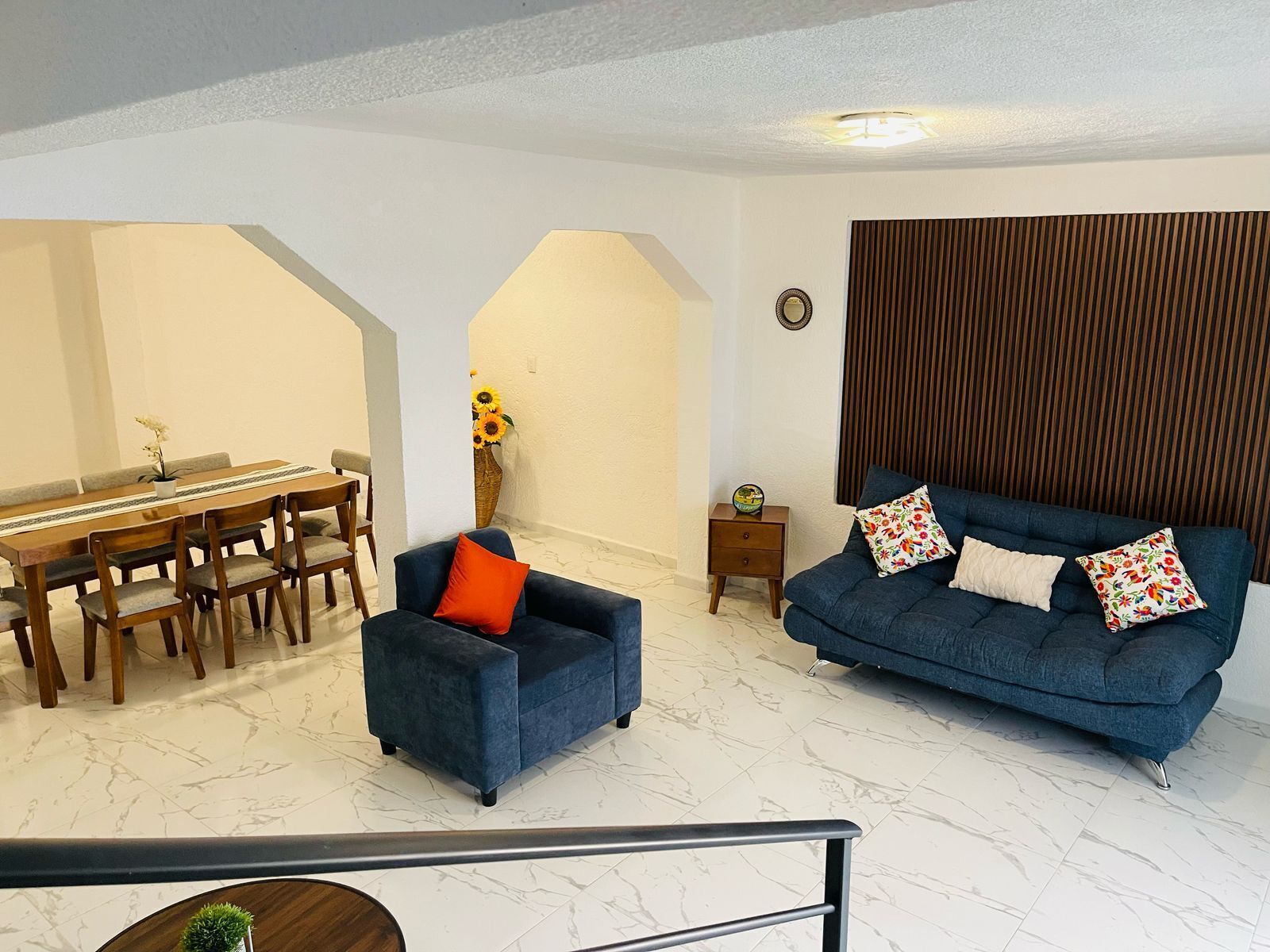 A living room with a dining area, a blue sofa with floral cushions, a small side table, and a vase with sunflowers.
