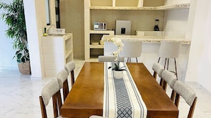 A dining area with a wooden table and six chairs, a kitchen in the background, and a potted plant. - Casa Mayor (Tenancingo)