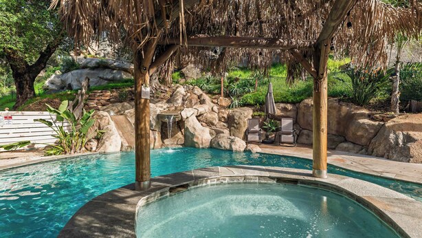 Uma área de piscina com hidromassagem, pérgula de madeira e paisagem rochosa.