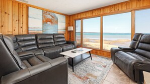 A living room with a leather sofa, a coffee table, and large windows overlooking a beach. - The Tides on East Bay (Traverse City)