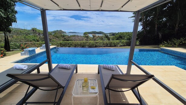 A poolside area with two lounge chairs, a glass table with a drink and a glass, and a view of a large pool and distant landscape. - Two-bedroom ocean view apartment with private pool near hotel facilities (Boca Chica)
