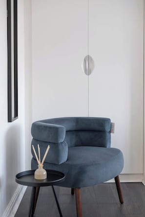 Quarto moderno com uma poltrona estofada azul escura, uma pequena mesa lateral preta e uma luminária fixada na parede.