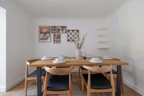 Uma sala de jantar com uma mesa de madeira posta para quatro pessoas, cadeiras estofadas pretas, um vaso com galhos e quadros nas paredes.