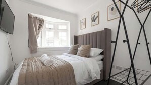 Ein Schlafzimmer mit Bett, einem fensterbelaubten Fenster, einem an der Wand befestigten Fernseher und einem Wäscheständer aus Metall.