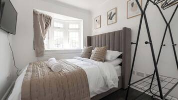A bedroom with a bed, a window with curtains, a wall-mounted TV, and a metal drying rack.