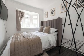 A bedroom with a bed, a window with curtains, a wall-mounted TV, and a metal drying rack. - Spacious 4BR Home I Sleeps 11 |Near Elizabeth Line (Romford)